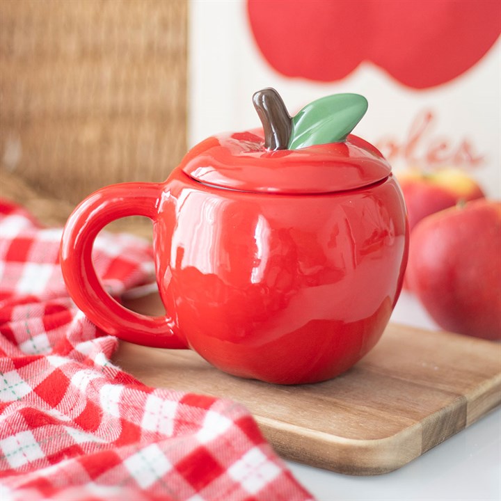 Red apple-shaped mug with a green leaf on a wooden cutting board with apples in the background.