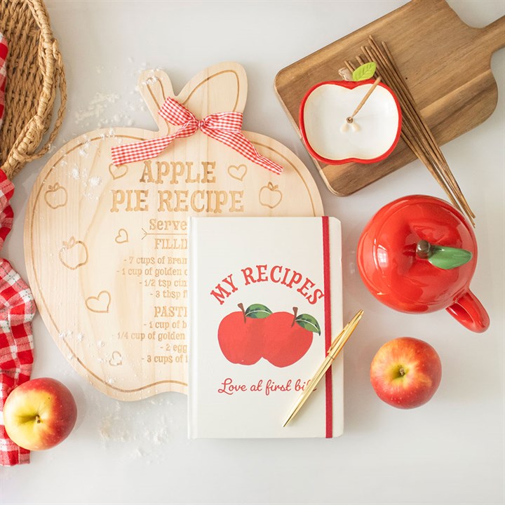 Apple-themed kitchen items including a pie recipe board, apples, a mug, and a notebook on a white surface.