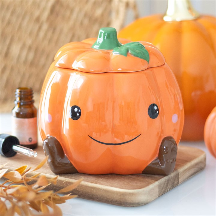 Ceramic pumpkin-shaped container with a face on a wooden board, with pumpkins and a bottle in the background.