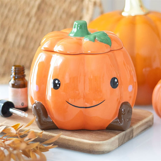 Ceramic pumpkin-shaped container with a face on a wooden board, with pumpkins and a bottle in the background.